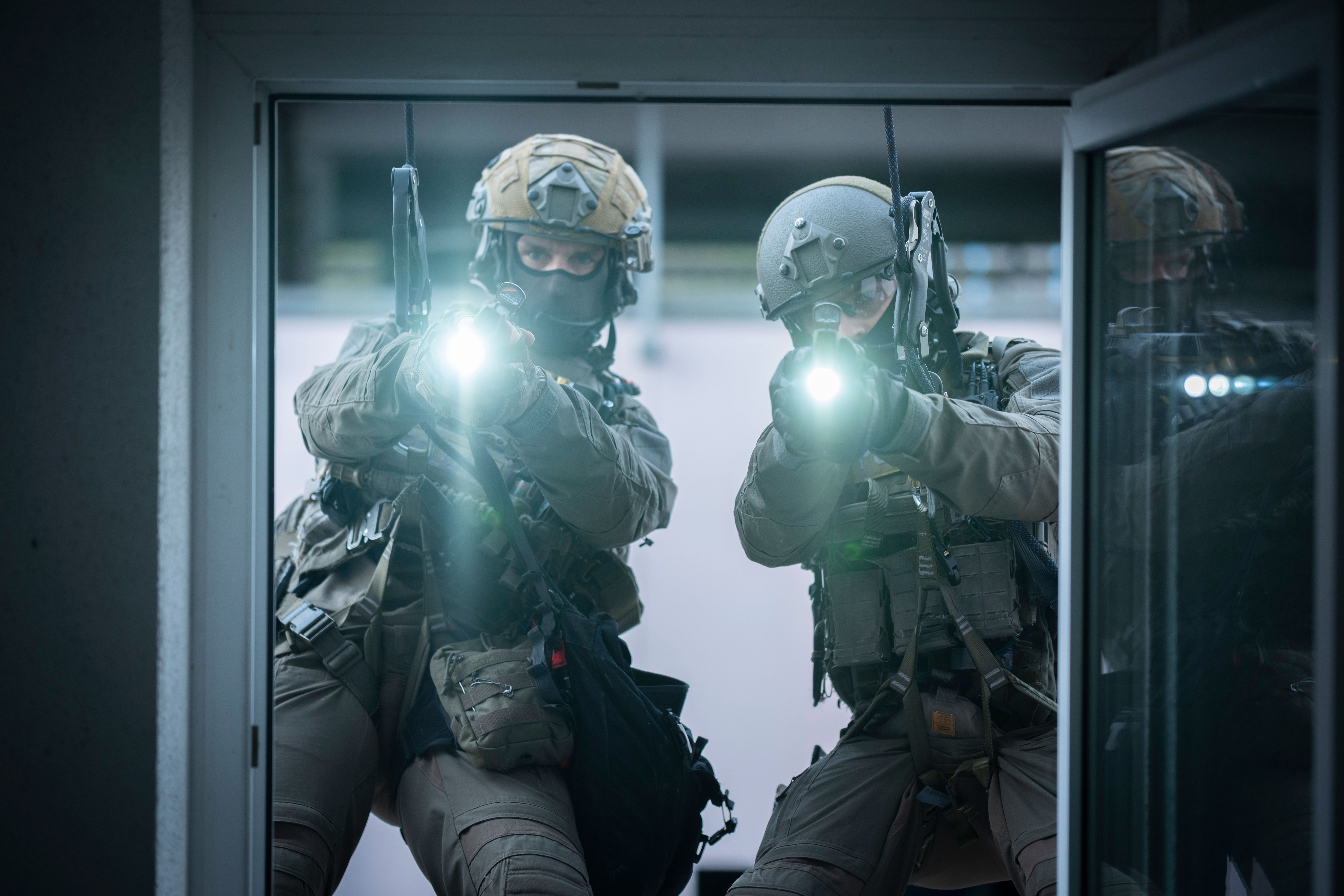 Two SEK officers abseil down to a window. They are wearing uniforms and helmets. They look through the window into the room and shine a light into it.