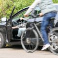 A man on a bicycle rides past a parked car whose driver is just opening her door. The police advise: Before opening a car door, always pay attention to the traffic behind you. The "Dutch grip" helps here.