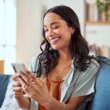 Eine Frau sieht auf einer Couch und schaut lächelnd ihr Handy an. Im Hintergrund sind weitere Möbel verschwommen zu sehen.