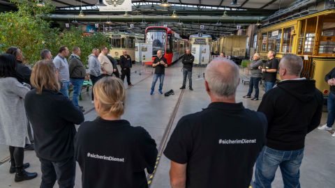 Es ist eine Halle zu sehen, in der Straßenbahnen gelagert sind. Im Vordergrund steht eine Gruppe Menschen im Kreis. Zwei Personen tragen T-Shirts mir der Aufschrift #Sicher im Dienst. Eine Person erklärt der Gruppe etwas.